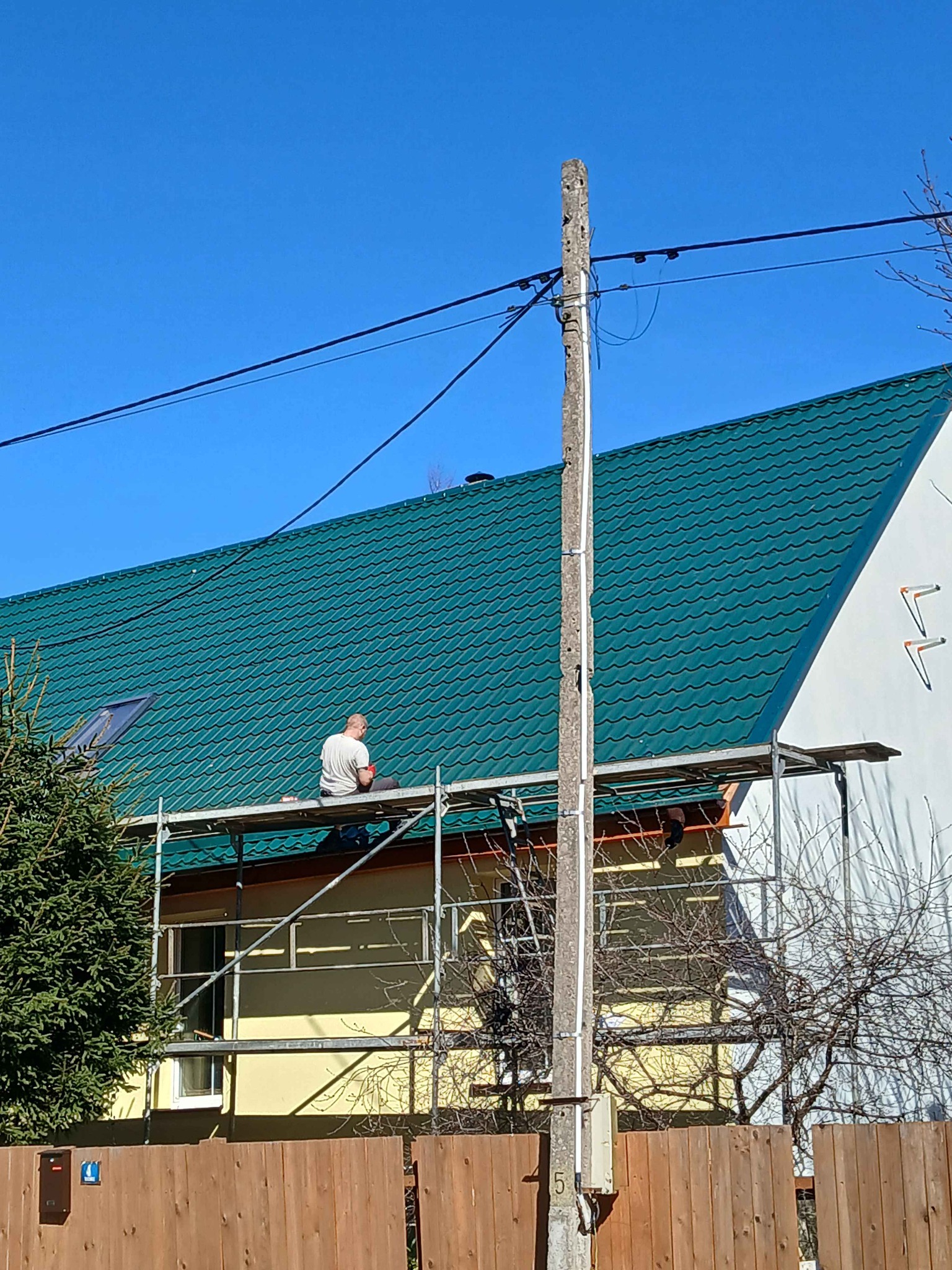 Praca na dachu z zielonej blachy falistej. Dekarz na rusztowaniu przy kominie, obok słup elektryczny. Widok na dom z drewnianym płotem.