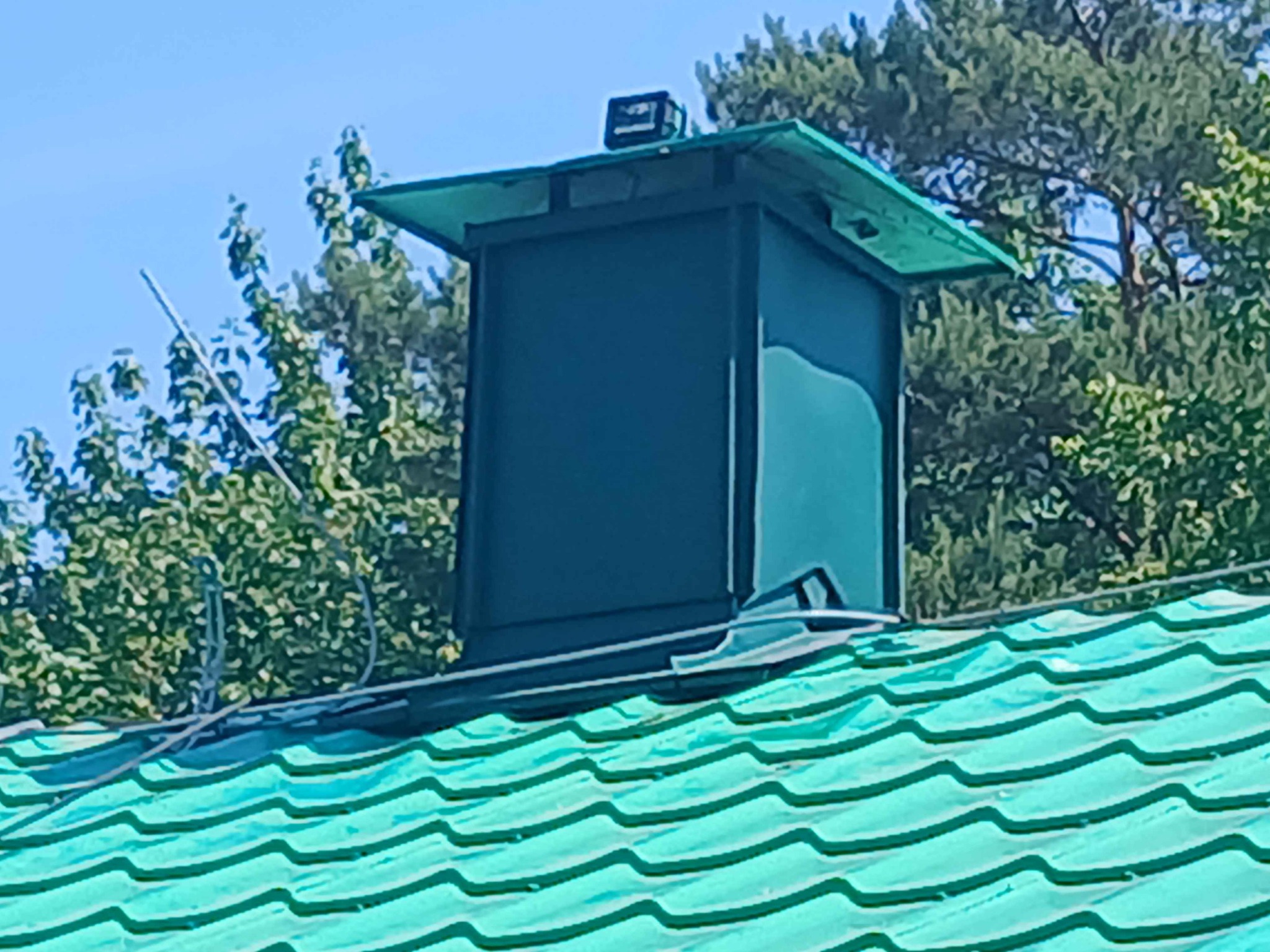 Szczegółowe ujęcie zielonego dachu z blachy falistej i ciemnego komina z metalową obudową oraz daszkiem, na tle zieleni drzew i błękitnego nieba.