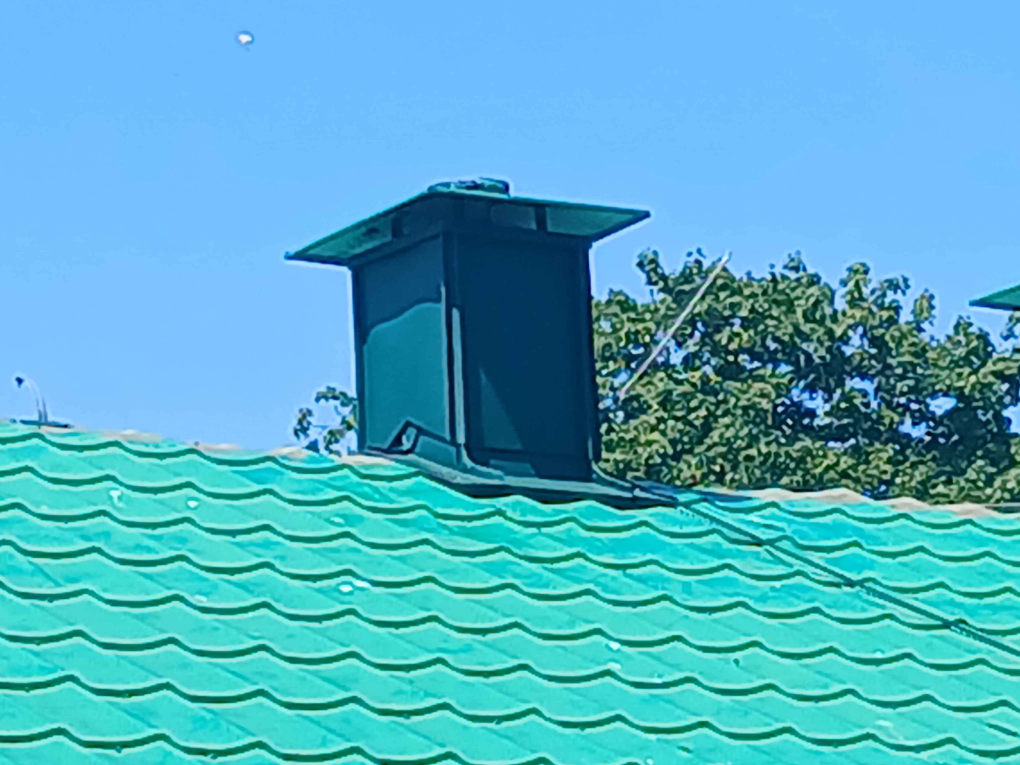 Zielony dach z blachy trapezowej i ciemny komin na tle błękitnego nieba i zieleni drzew. Komin z metalową czapą. Widok z dołu.