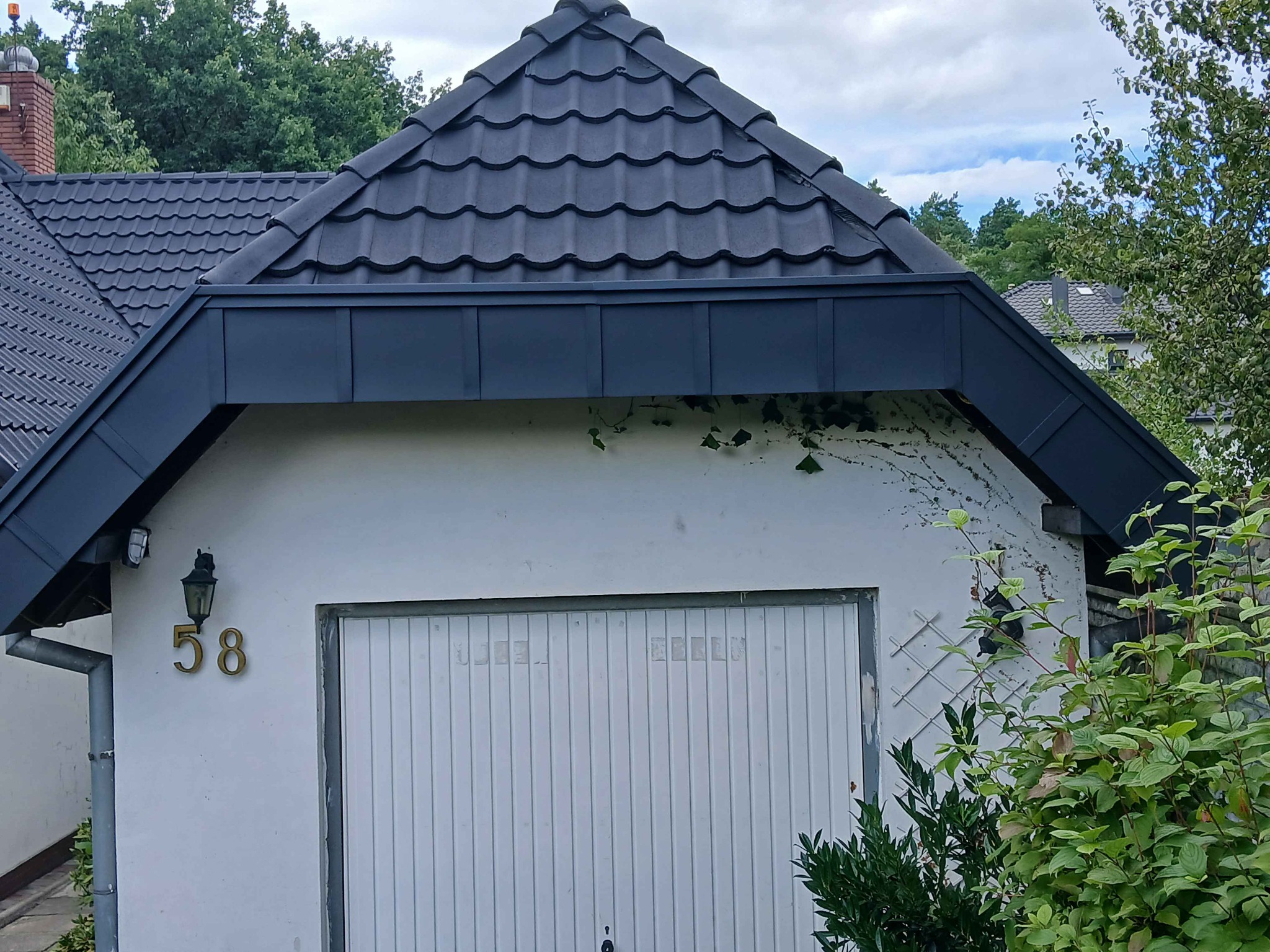 Ciemna dachówka ceramiczna na garażu z numerem 58, obróbki blacharskie w kolorze grafitowym. Widoczne fragmenty zieleni i błękitne niebo.