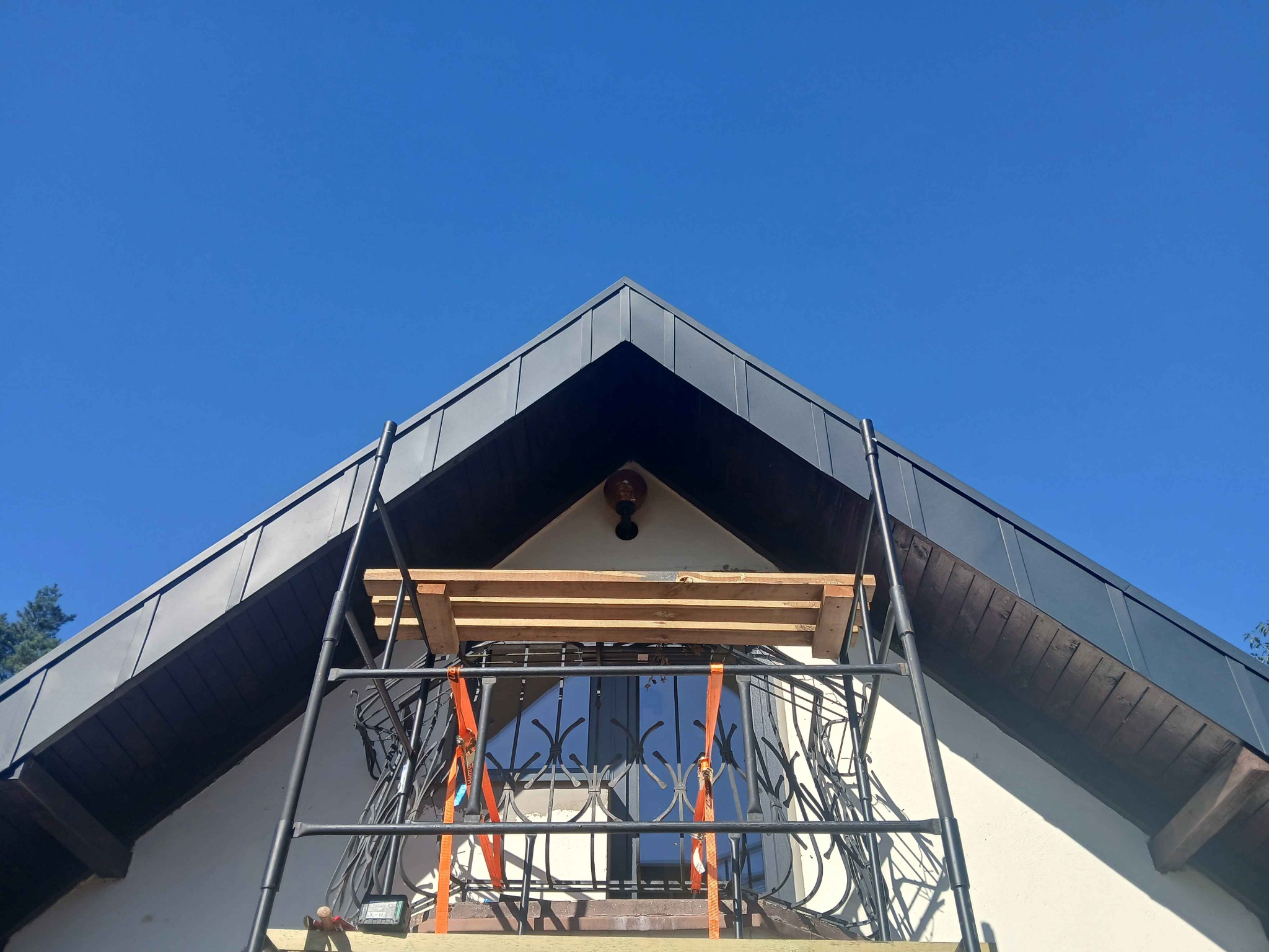 Szary dach z blachy na budynku z balkonem, widok z dołu. Na balkonie rusztowanie z deskami i pasami mocującymi. Błękitne niebo w tle.