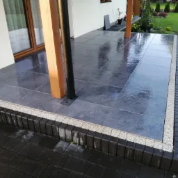 Błyszczący, ciemnoszary taras z płytek ceramicznych, otoczony obrzeżem z jasnego grysu i kostki brukowej, z drewnianymi słupami podtrzymującymi zadaszenie, widok na zadbany ogród.