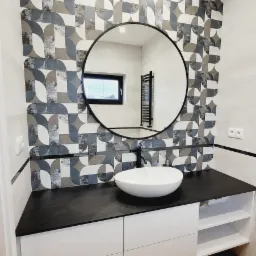 Nowoczesna łazienka z okrągłym lustrem, umywalką nablatową i geometrycznymi płytkami na ścianie, widok na grzejnik drabinkowy w odbiciu lustra.