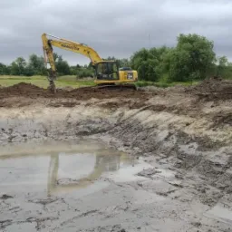 Żółta koparka Komatsu 210 LC pogłębia wykop wypełniony wodą na tle zieleni i pochmurnego nieba.