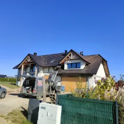 Nowy dom w trakcie budowy z częściowo ukończonym dachem z ciemnej dachówki, widoczny dekarz na drabinie i samochód dostawczy z napisem 'Pokrycia dachowe' na tle błękitnego nieba.