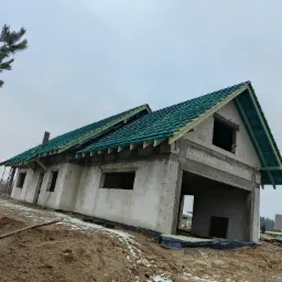 Surowy stan domu z częściowo ukończonym dachem z zielonej blachy, widoczne pustaki i teren wokół w trakcie prac ziemnych.