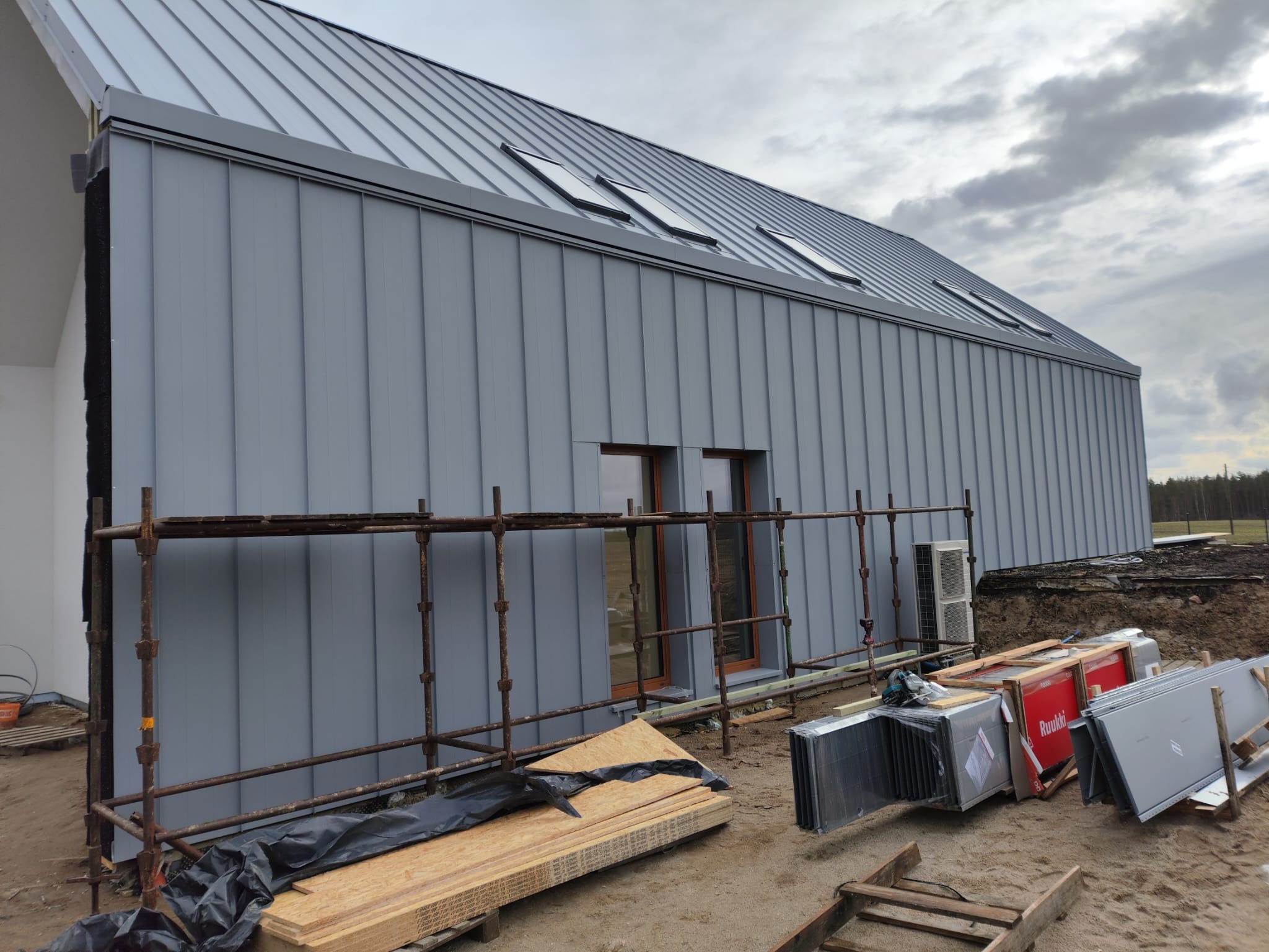 Nowoczesny dom z szarą blachą na elewacji i dachu, widoczne okna dachowe, rusztowanie przy ścianie, materiały budowlane na ziemi. Ujęcie z dołu, pochmurne niebo.