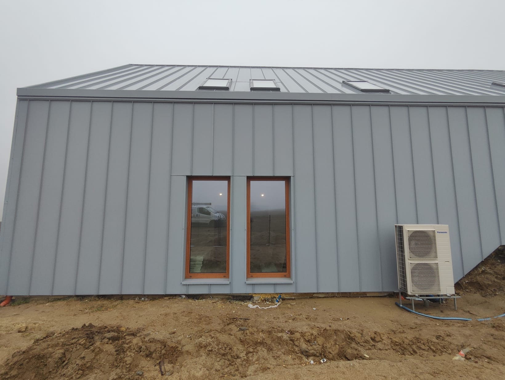 Szara elewacja budynku z blachy trapezowej, widoczne dwa okna z brązowymi ramami i klimatyzator na zewnątrz. Dach z trzema oknami dachowymi.