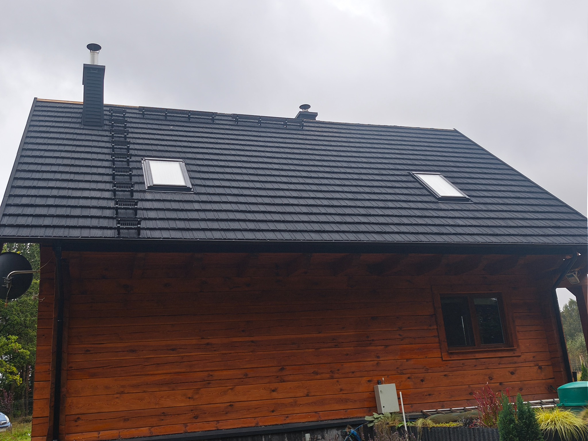 Czarny dach z blachy trapezowej z oknami dachowymi i drabinką kominiarską na drewnianym domu jednorodzinnym w pochmurny dzień. Widok z dołu.