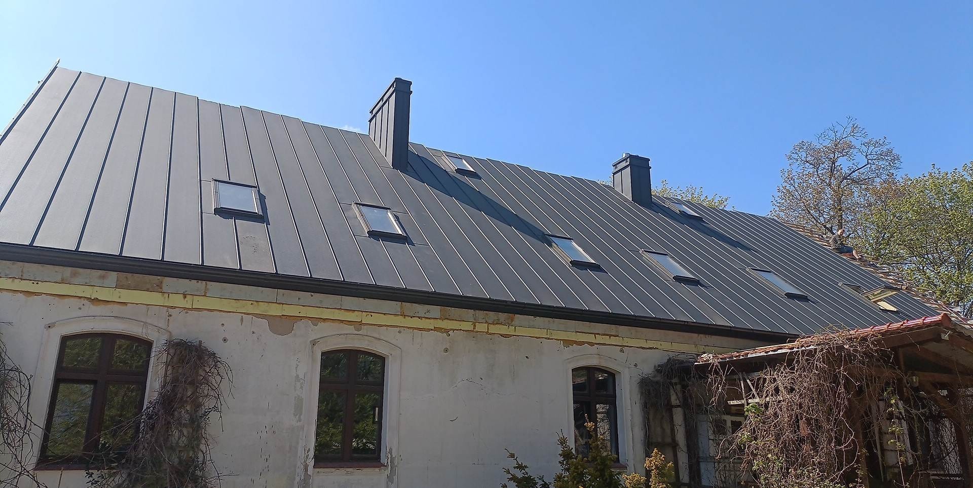 Szary, blaszany dach z oknami dachowymi na budynku z jasną elewacją i ciemnymi oknami. Widoczne kominy i fragment zarośniętej roślinnością werandy.