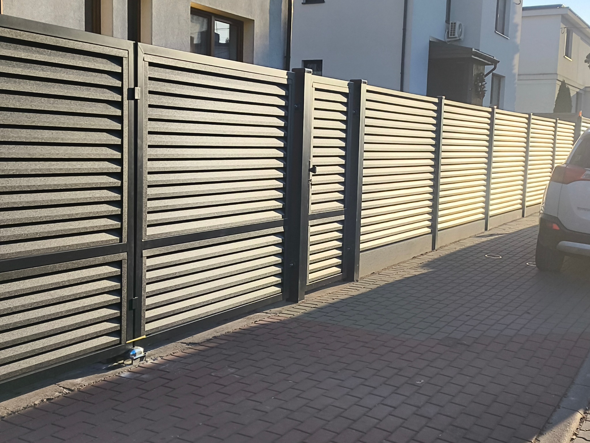 Nowoczesne ogrodzenie posesji z poziomych paneli w kolorze szaro-beżowym, z przesuwną bramą wjazdową i furtką, widok z kostki brukowej.