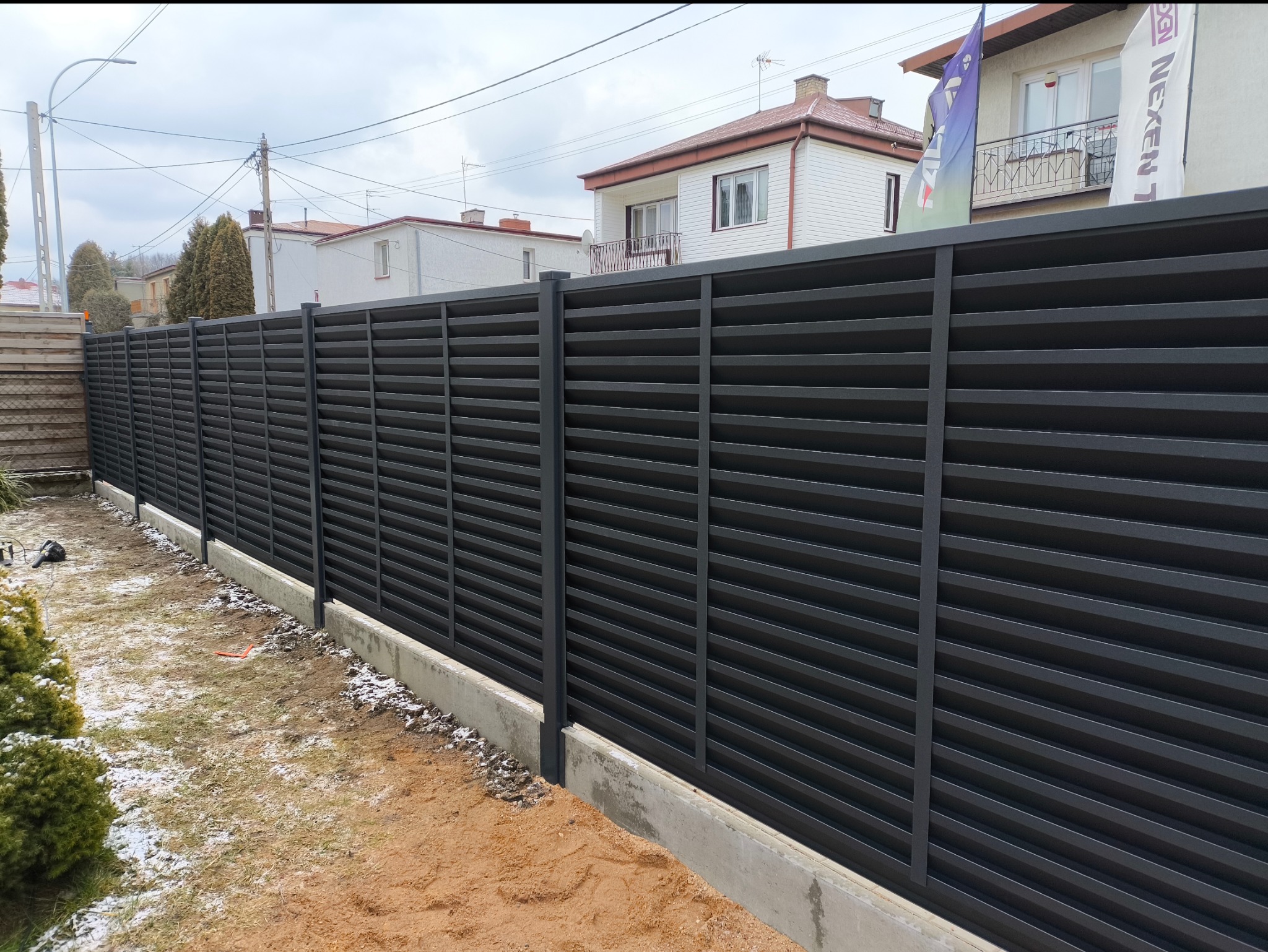 Nowoczesne, poziome, grafitowe ogrodzenie panelowe na betonowej podmurówce, widok z zewnątrz posesji, z elementami trawnika i śniegu.