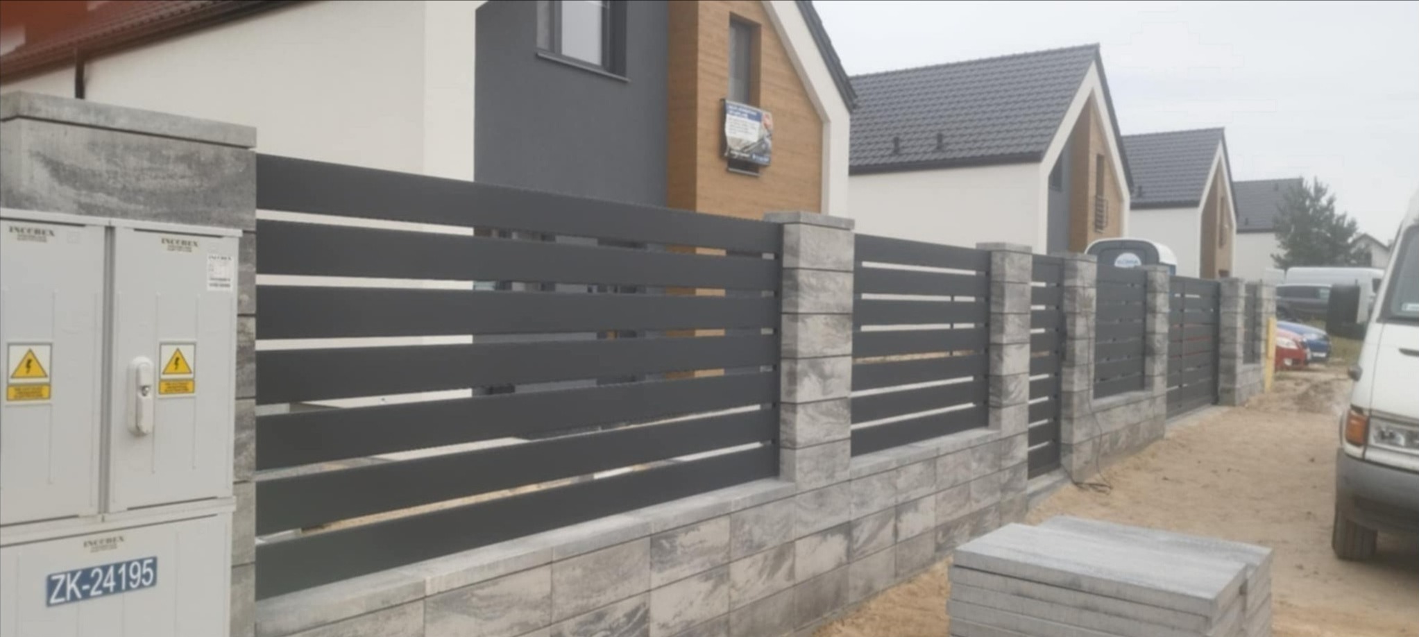 Nowoczesne ogrodzenie panelowe z poziomych, szarych profili, osadzone na słupkach i podmurówce z szarej kostki imitującej kamień, na tle nowo wybudowanych domów.