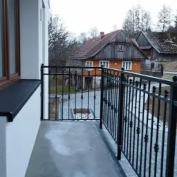 Barierka balkonowa z czarnych, metalowych prętów z ozdobnymi kulkami, widok na wiejską zabudowę i drogę.