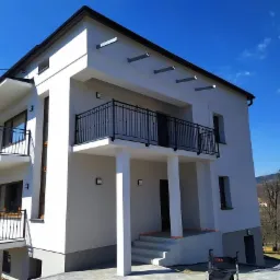 Nowoczesny, dwupiętrowy dom z balkonami zabezpieczonymi czarnymi, metalowymi balustradami, widok z dołu na tle błękitnego nieba.