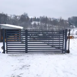 Nowoczesna, pozioma brama wjazdowa w kolorze antracytowym, widziana zimą na tle zaśnieżonego krajobrazu z budynkami i drzewami.