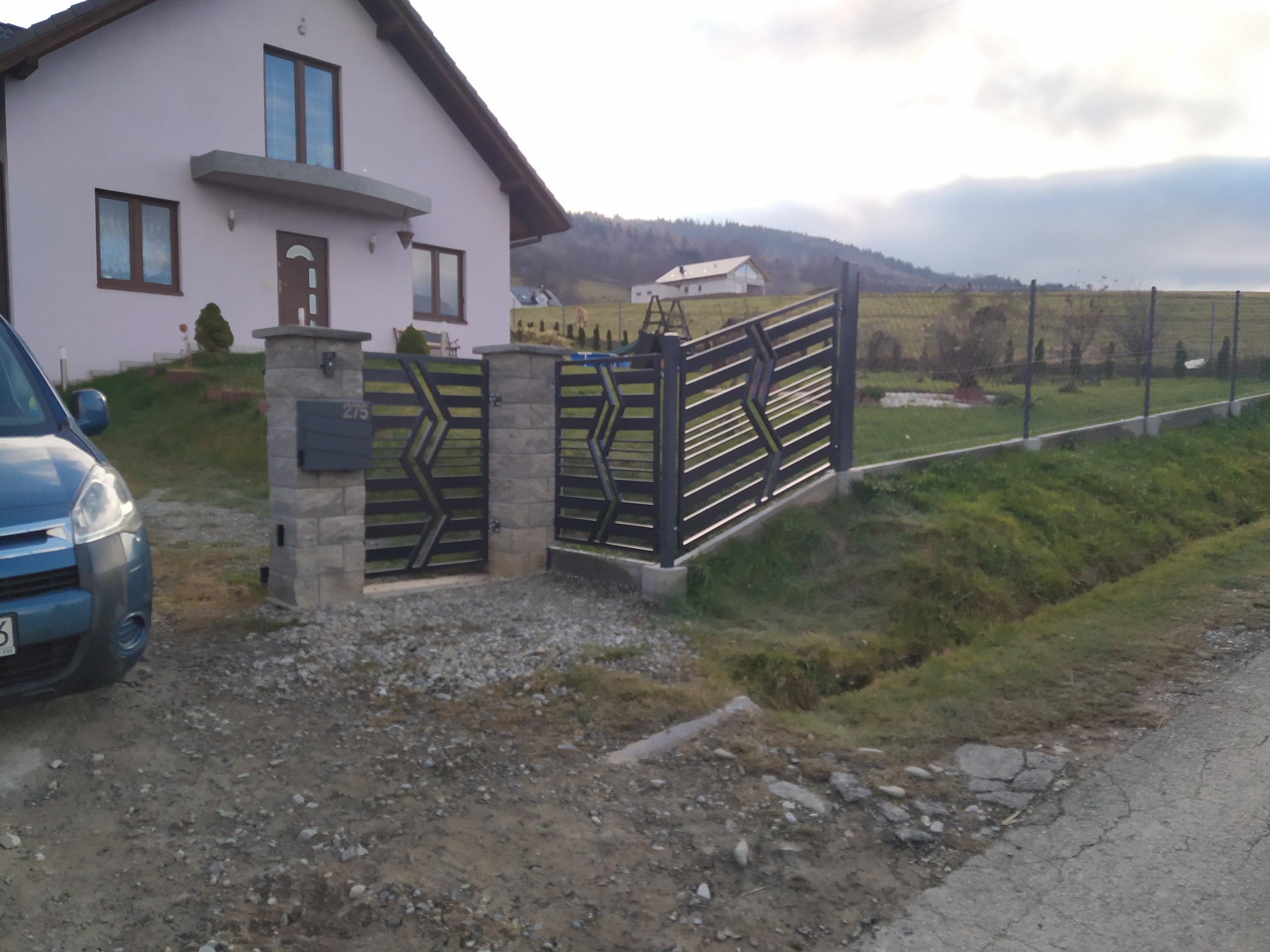 Nowoczesna, czarna brama wjazdowa z geometrycznym wzorem, zamontowana na słupkach z szarej kostki, na tle domu w odcieniach różu i krajobrazu z lekkim zachmurzeniem.