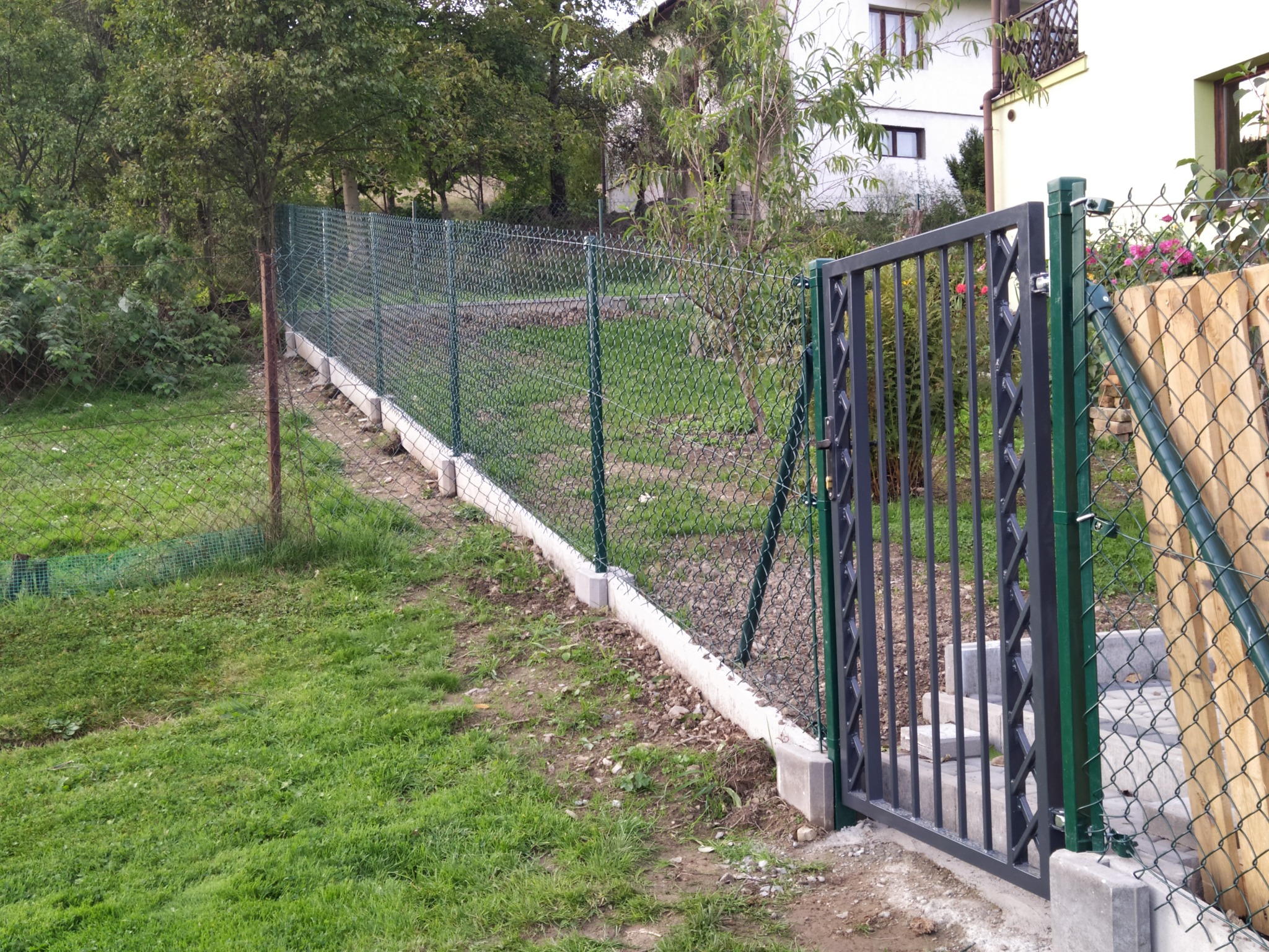 Zielona siatka ogrodzeniowa z podmurówką betonową i furtką z metalowymi prętami na tle domu i zieleni.