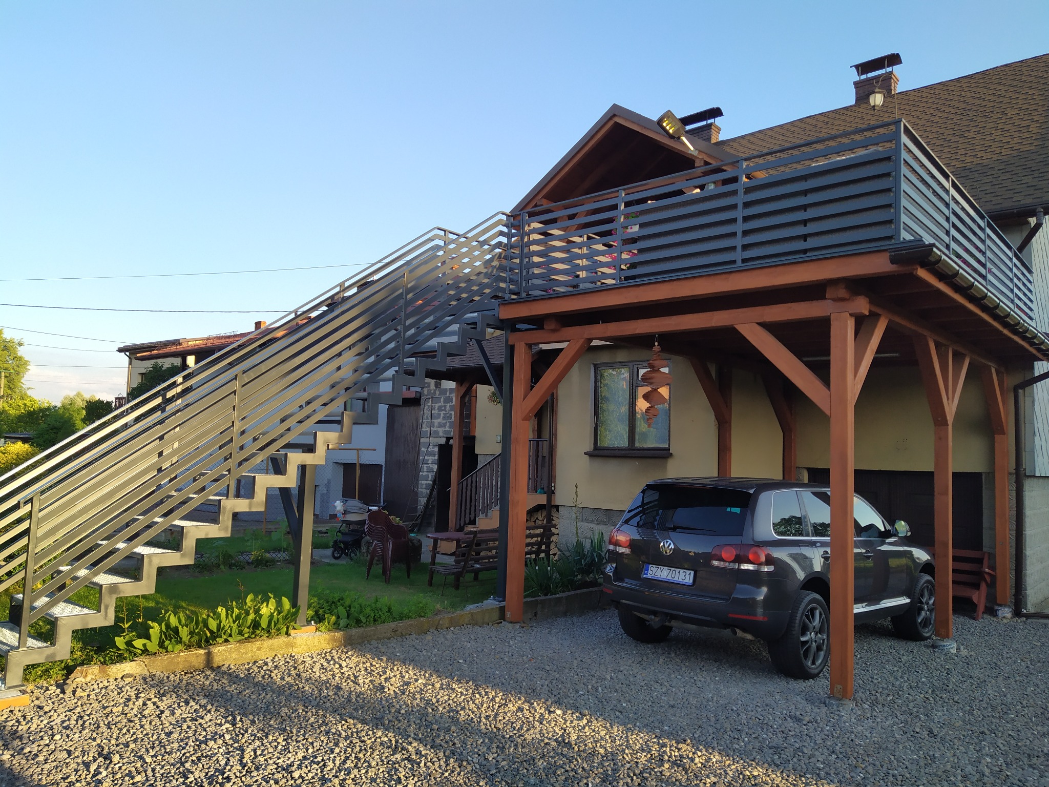 Zewnętrzne schody metalowe prowadzące na taras z metalową balustradą, obok zaparkowany ciemny samochód osobowy.