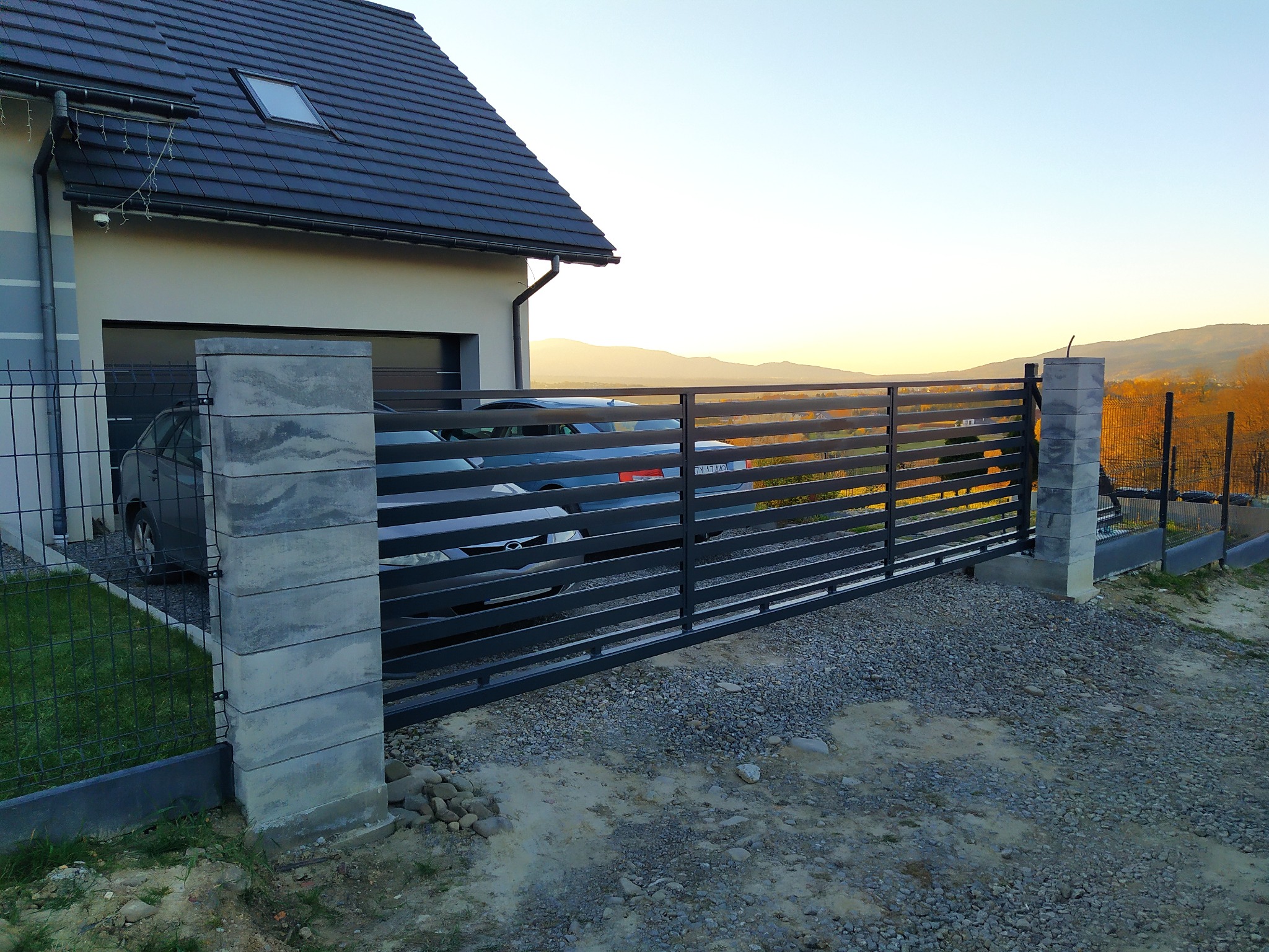 Nowoczesna, pozioma brama przesuwna z metalowymi profilami, szara, z widokiem na góry w oddali, zamontowana przy domu z garażem i ogrodzeniem panelowym.