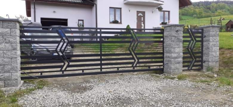 Nowoczesna, pozioma, czarna konstrukcja z metalu z motywem zygzaków, osadzona na słupkach z szarej kostki, na tle domu z jasną elewacją i zielonego wzgórza.