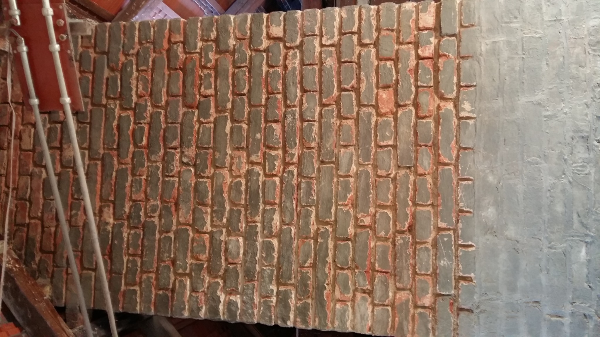 Fragment naprawianego komina od strony strychu, z widocznym przemurowaniem ponad kalenicą, cegły z szarą zaprawą i czerwonymi śladami, część komina obłożona szarym tynkiem.