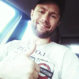 Uśmiechnięty hydraulik w czapce, robiący selfie w samochodzie, z kciukiem w górę, ubrany w koszulkę z motywem London Underground.