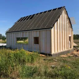 Surowy szkielet drewnianego domu z częściowo ułożonymi łatami dachowymi na tle błękitnego nieba i zarośniętej trawy.