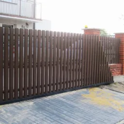 Brązowa brama przesuwna z pionowych paneli, zamontowana na posesji z ceglanymi słupkami i kostką brukową na podjeździe.