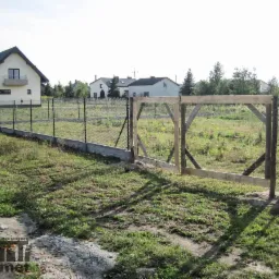 Ogrodzenie z siatki ocynkowanej z podmurówką betonową, widoczna brama w trakcie montażu z surowego drewna, w tle dom jednorodzinny i zarośnięta działka.