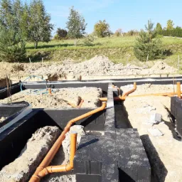 Fundamenty domu w trakcie budowy z widocznymi pomarańczowymi rurami kanalizacyjnymi ułożonymi w wykopach, zabezpieczone czarną izolacją przeciwwilgociową.