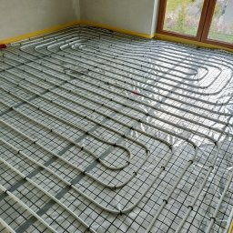 INSTALFIX - Białe rurki instalacji ogrzewania podłogowego ułożone na folii z siatką, widoczne okno z zielonym ogrodem na zewnątrz.