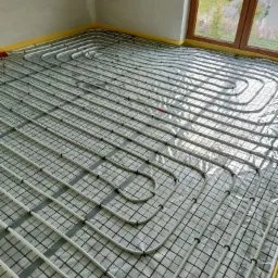 Białe rurki instalacji ogrzewania podłogowego ułożone na folii z siatką, widoczne okno z zielonym ogrodem na zewnątrz.