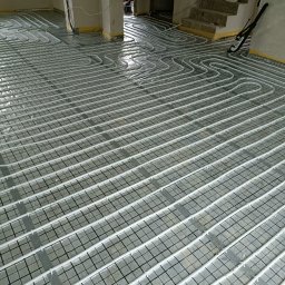 INSTALFIX - Białe rury ogrzewania podłogowego ułożone na siatce w surowym pomieszczeniu, widoczne żółte taśmy zabezpieczające ściany, schody w tle.