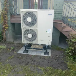 Zewnętrzna jednostka pompy ciepła Hitachi, umieszczona na specjalnych wspornikach na płytkach chodnikowych, przed budynkiem z zielonej cegły i metalową balustradą.