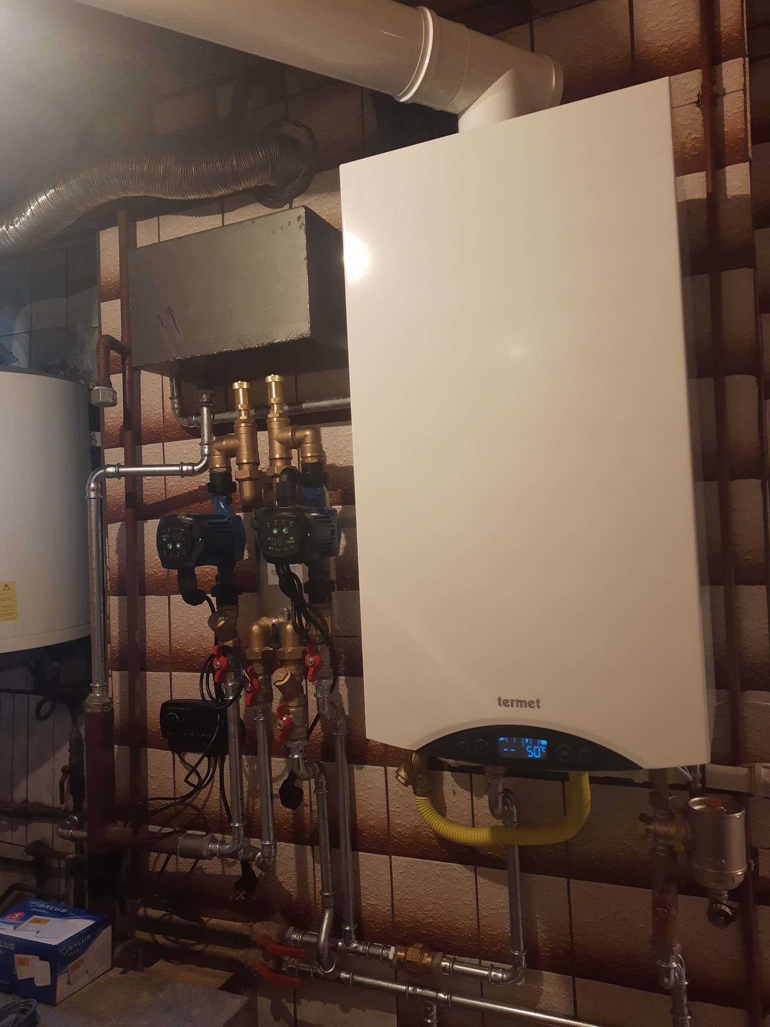 Biały piec gazowy marki Termet zamontowany na ścianie z brązowych płytek, widoczne rury, zawory, pompy obiegowe i elastyczny przewód gazowy.