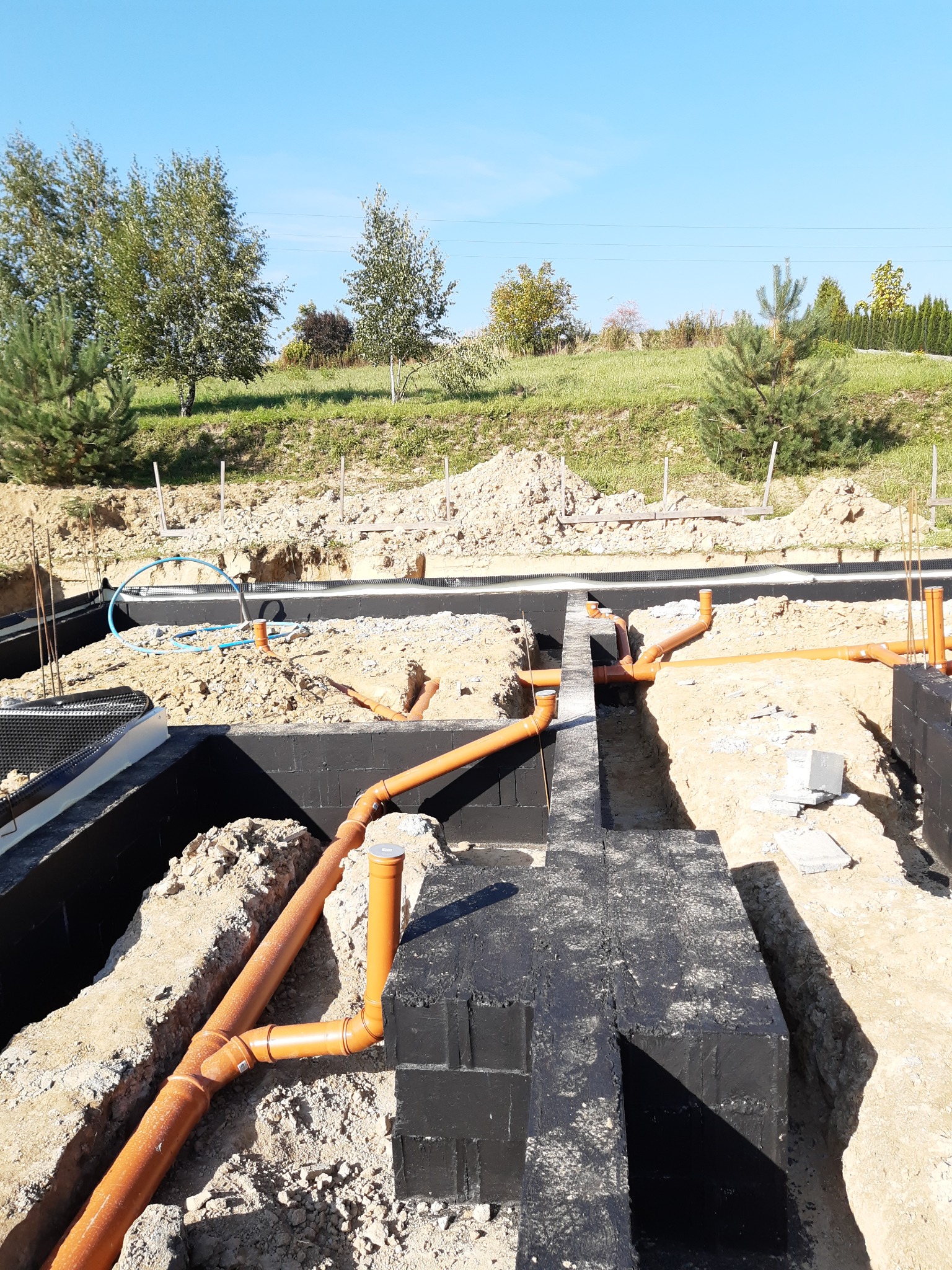 Fundamenty domu w trakcie budowy z widocznymi pomarańczowymi rurami kanalizacyjnymi ułożonymi w wykopach, zabezpieczone czarną izolacją przeciwwilgociową.