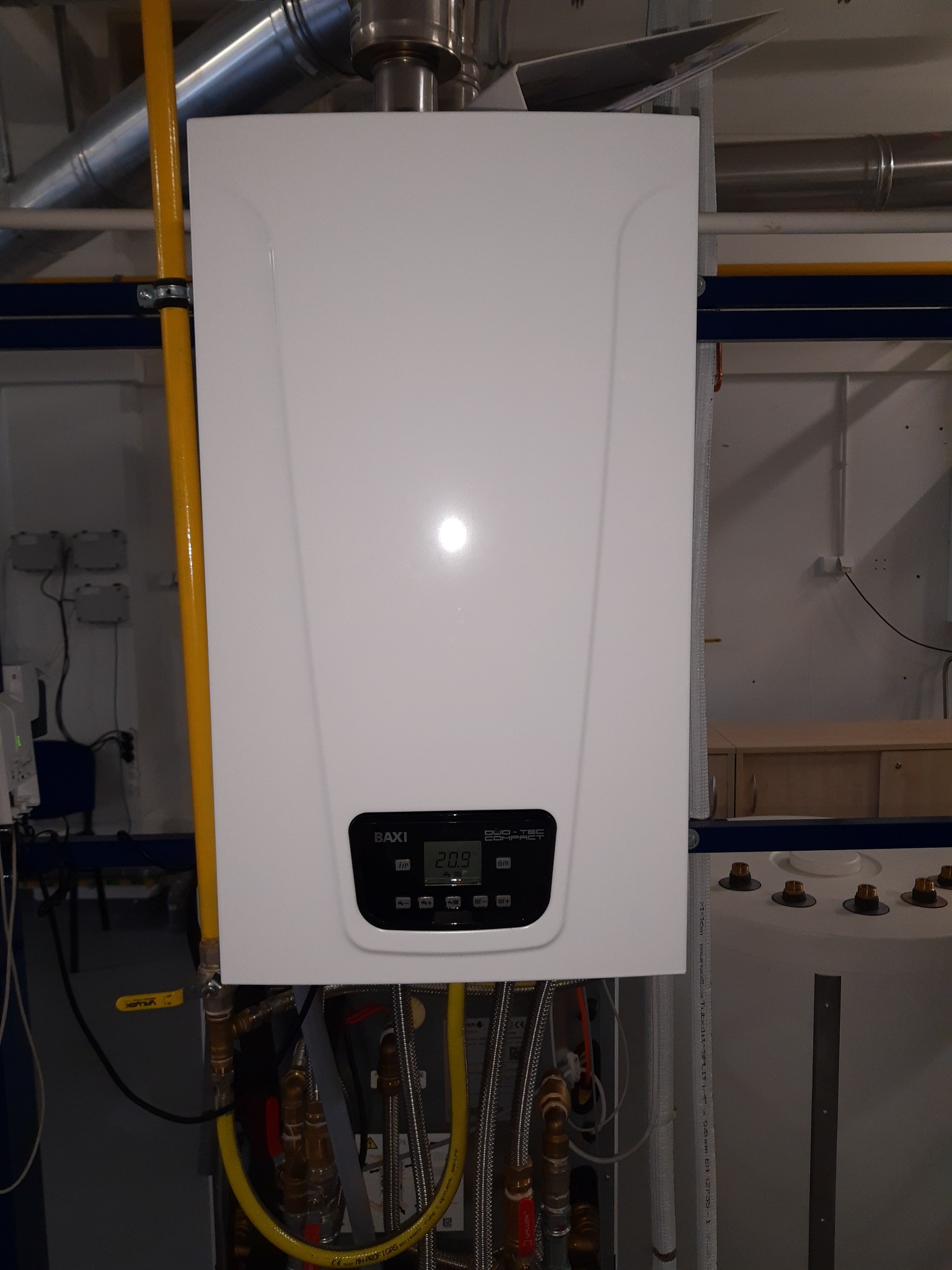 Biały piec gazowy marki Baxi Duo-Tec Compact zamontowany w pomieszczeniu technicznym, widoczne żółte rury gazowe i metalowe przewody instalacyjne.