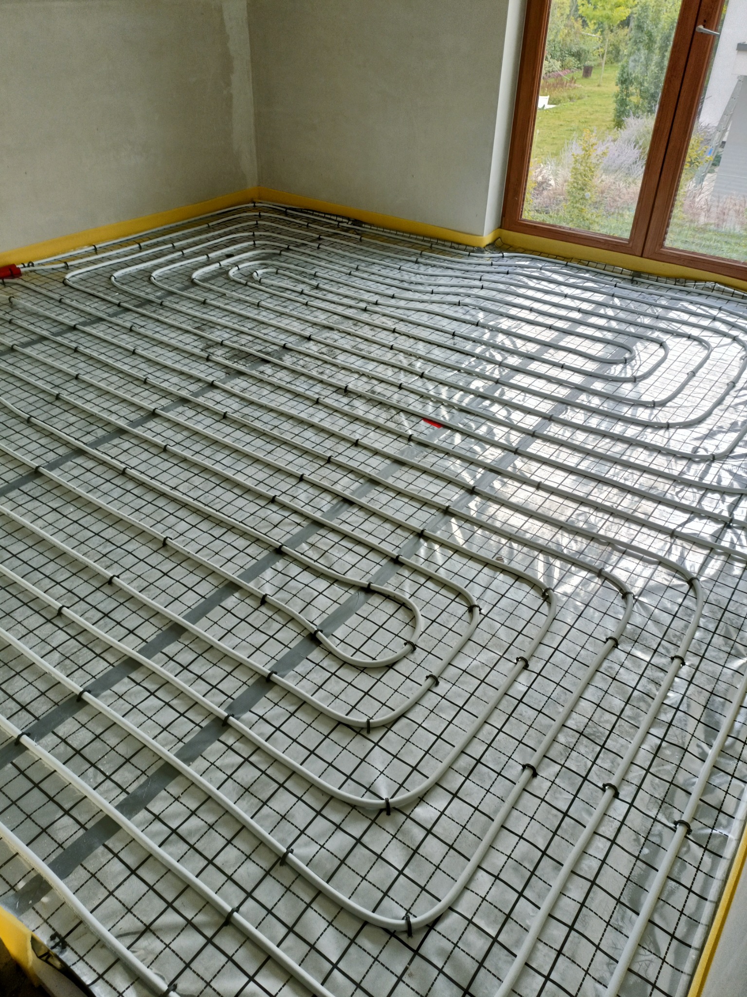 Białe rurki instalacji ogrzewania podłogowego ułożone na folii z siatką, widoczne okno z zielonym ogrodem na zewnątrz.