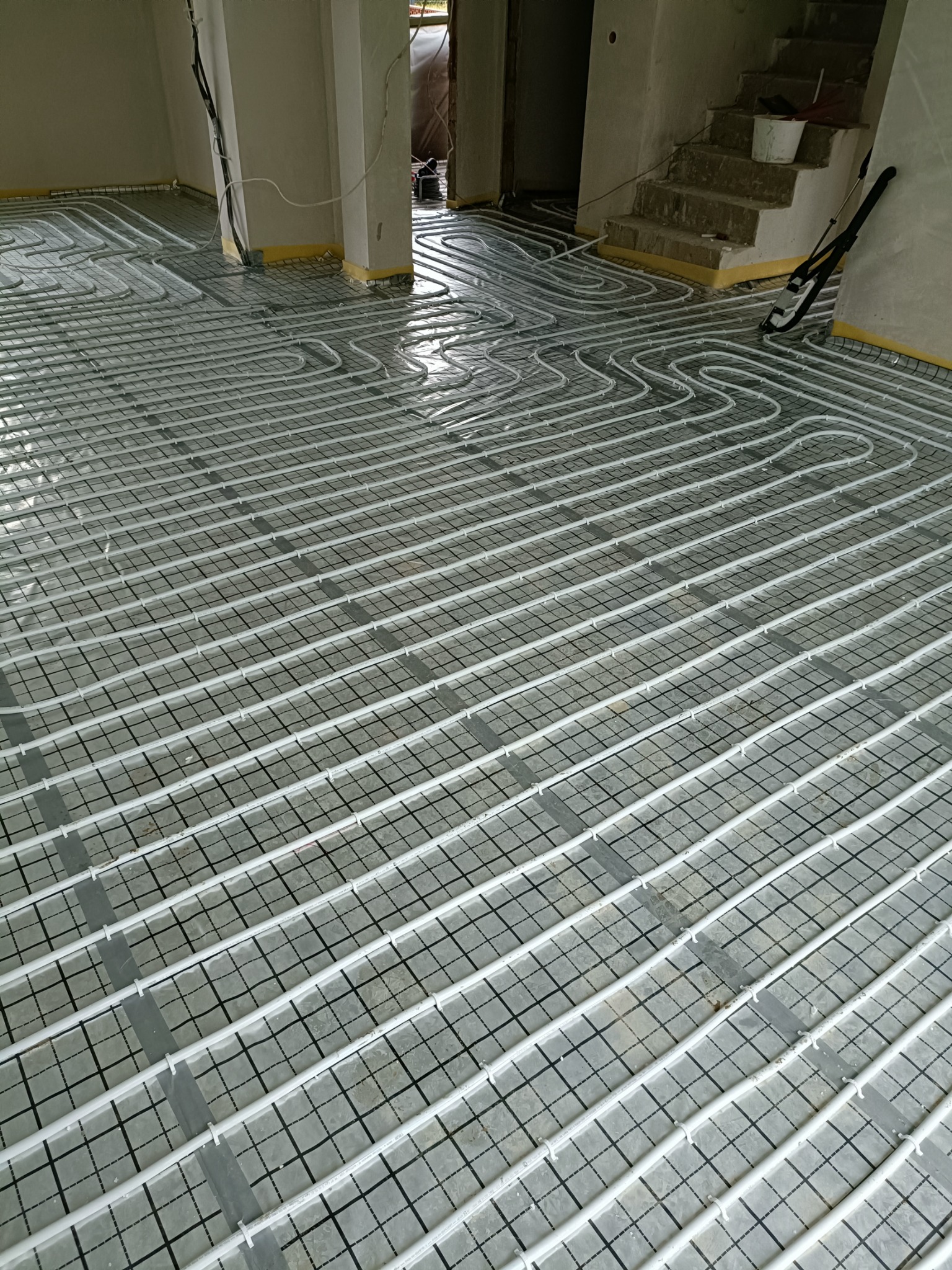 Białe rury ogrzewania podłogowego ułożone na siatce w surowym pomieszczeniu, widoczne żółte taśmy zabezpieczające ściany, schody w tle.