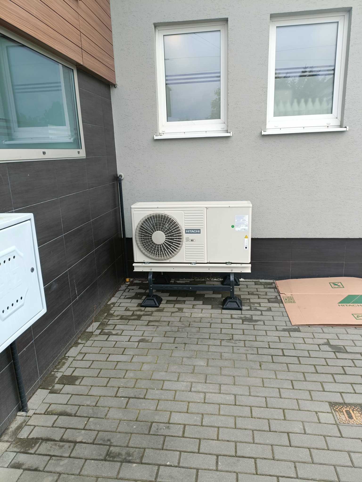 Zewnętrzna jednostka pompy ciepła Hitachi zamontowana na metalowej konstrukcji, umieszczona na brukowanej powierzchni przed budynkiem z szarą elewacją i dwoma oknami.