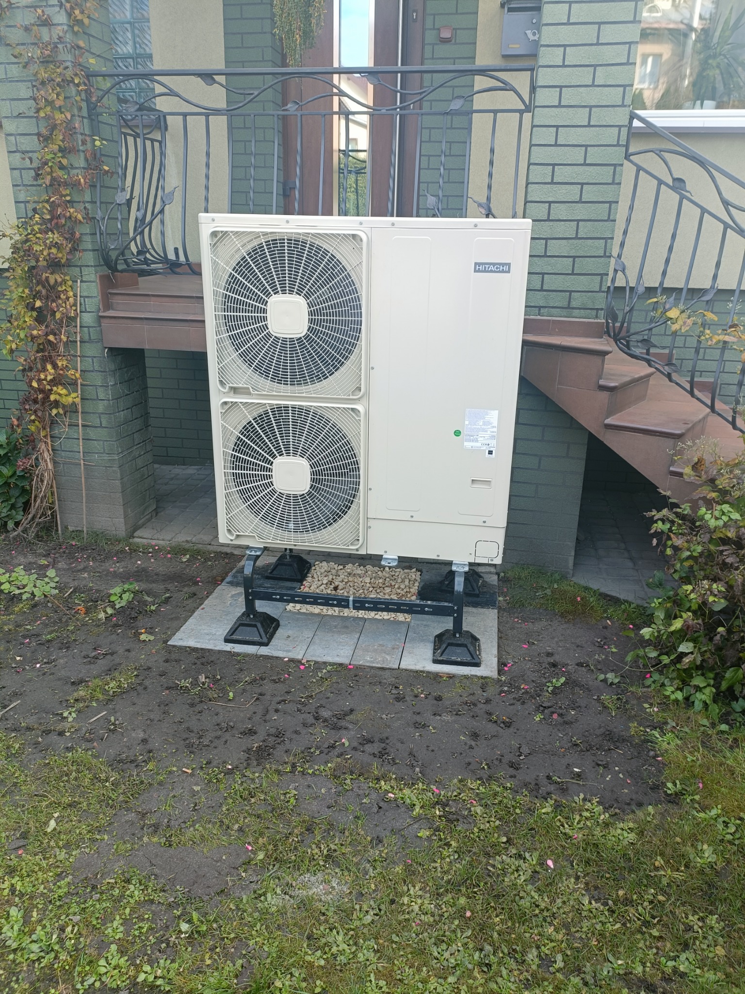 Zewnętrzna jednostka pompy ciepła Hitachi, umieszczona na specjalnych wspornikach na płytkach chodnikowych, przed budynkiem z zielonej cegły i metalową balustradą.