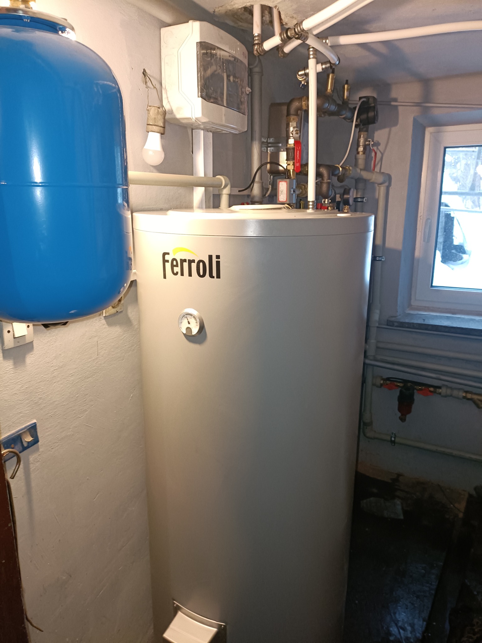 Szary, cylindryczny zasobnik ciepłej wody marki Ferroli z termometrem, obok niebieski zbiornik wyrównawczy, widoczne rury i zawory instalacji w piwnicy.