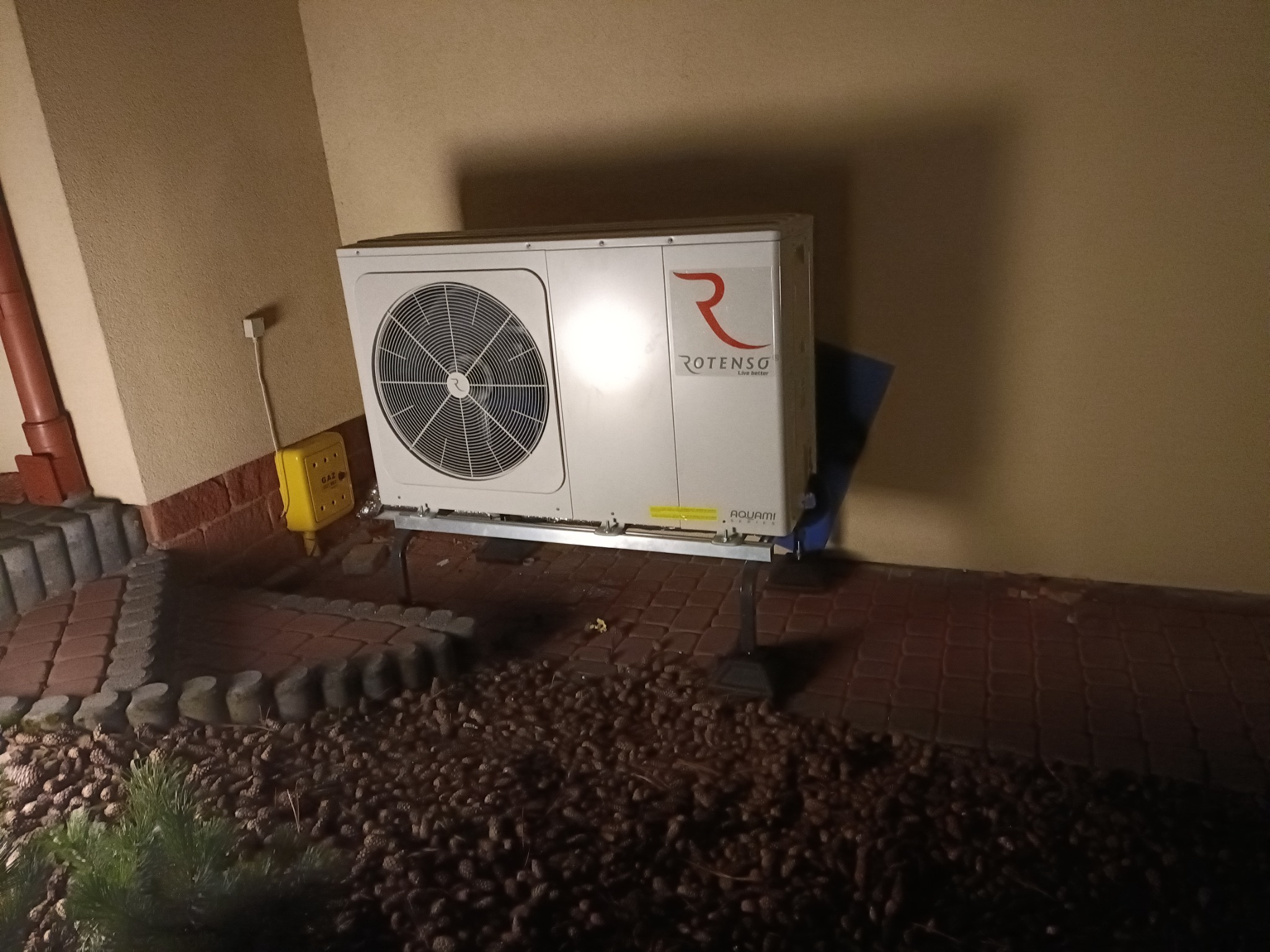 Biała pompa ciepła marki Rotenso zamontowana na zewnątrz budynku, widoczna instalacja gazowa w żółtej obudowie.