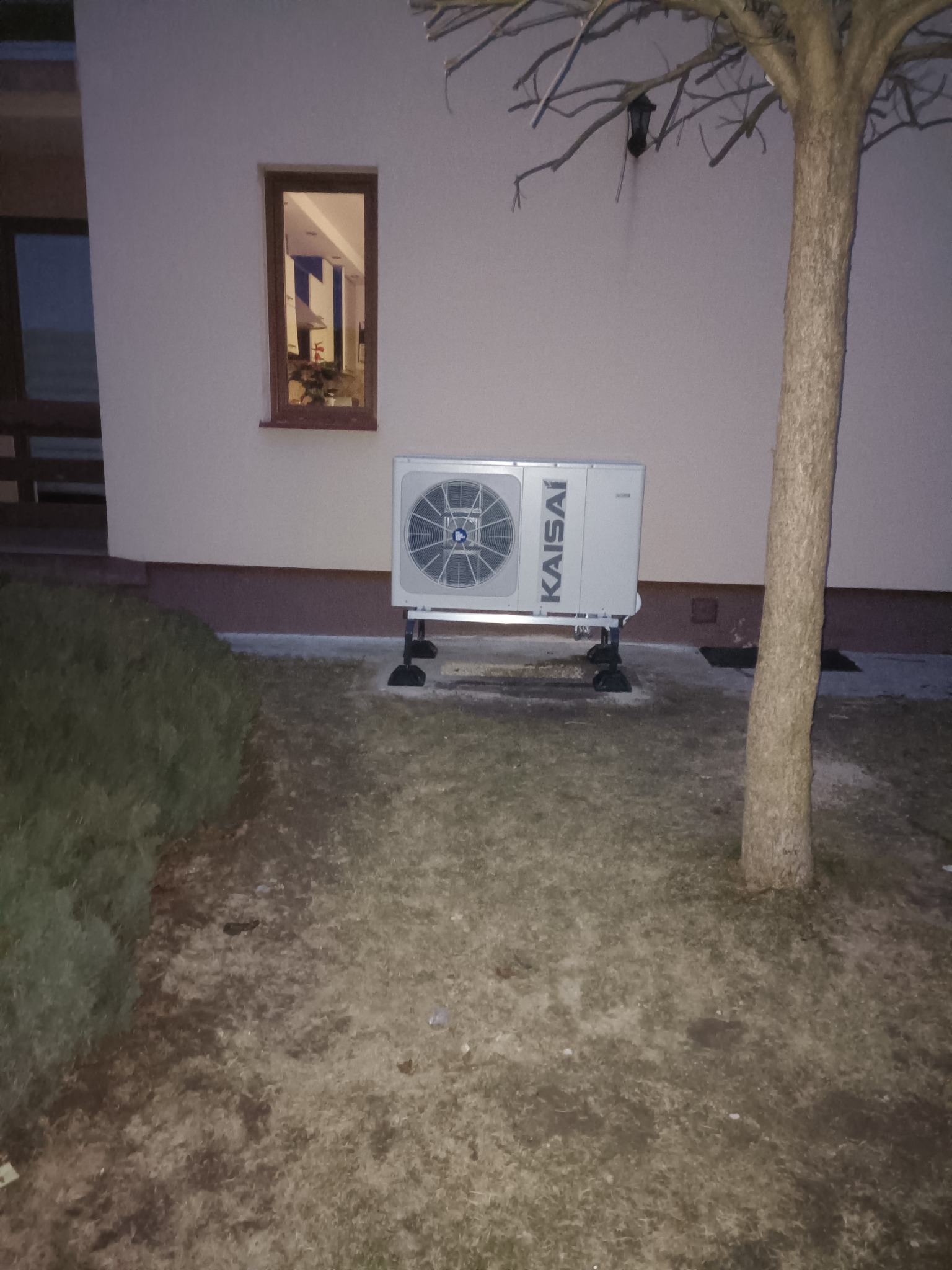 Biała pompa ciepła marki Kaisai zamontowana na zewnątrz budynku, widoczna z perspektywy trawnika z drzewem bez liści, z oknem budynku w tle.