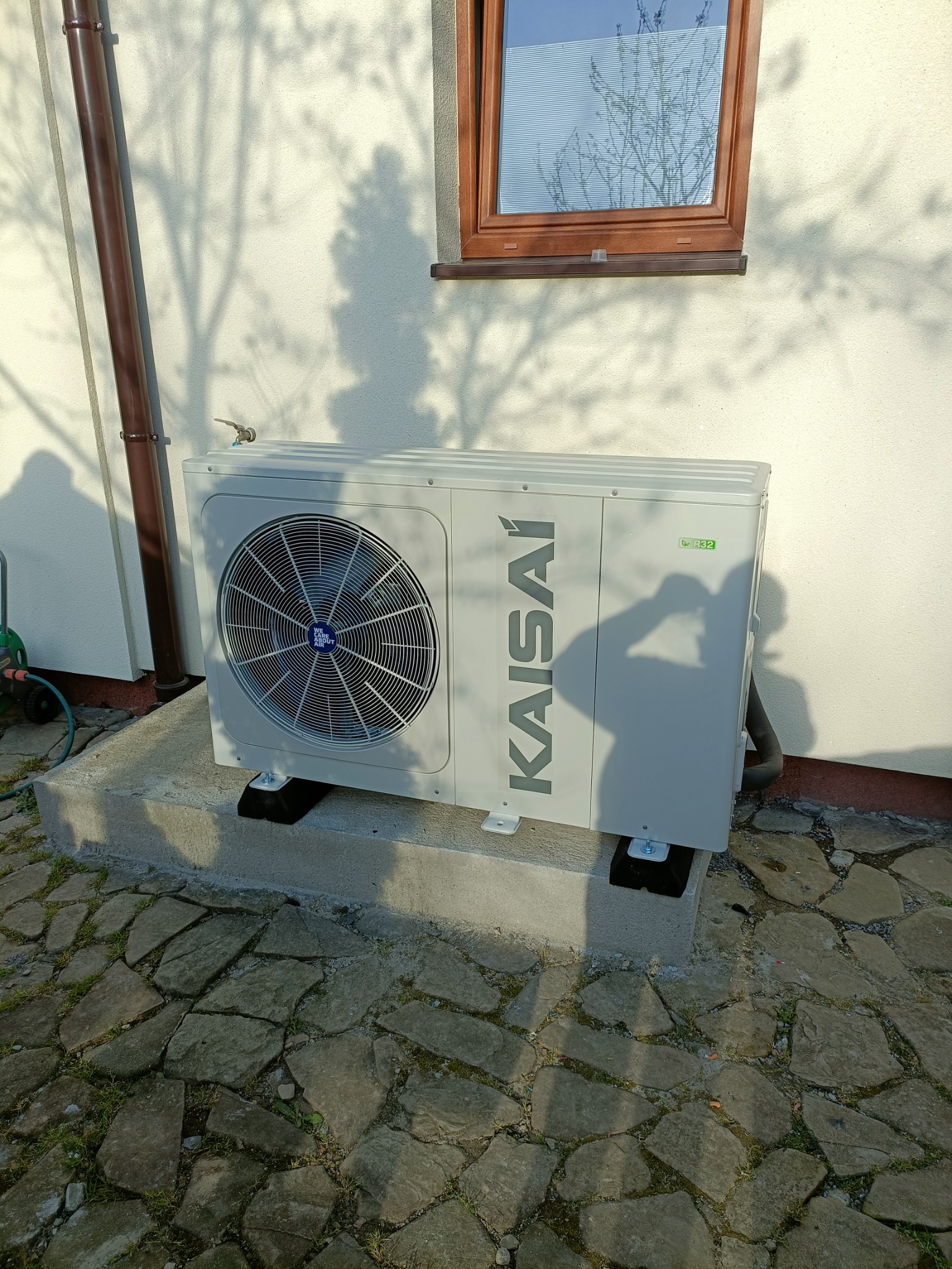 Zewnętrzna jednostka klimatyzacji Kaisai zamontowana na betonowym podeście przed budynkiem, cień osoby na ścianie.