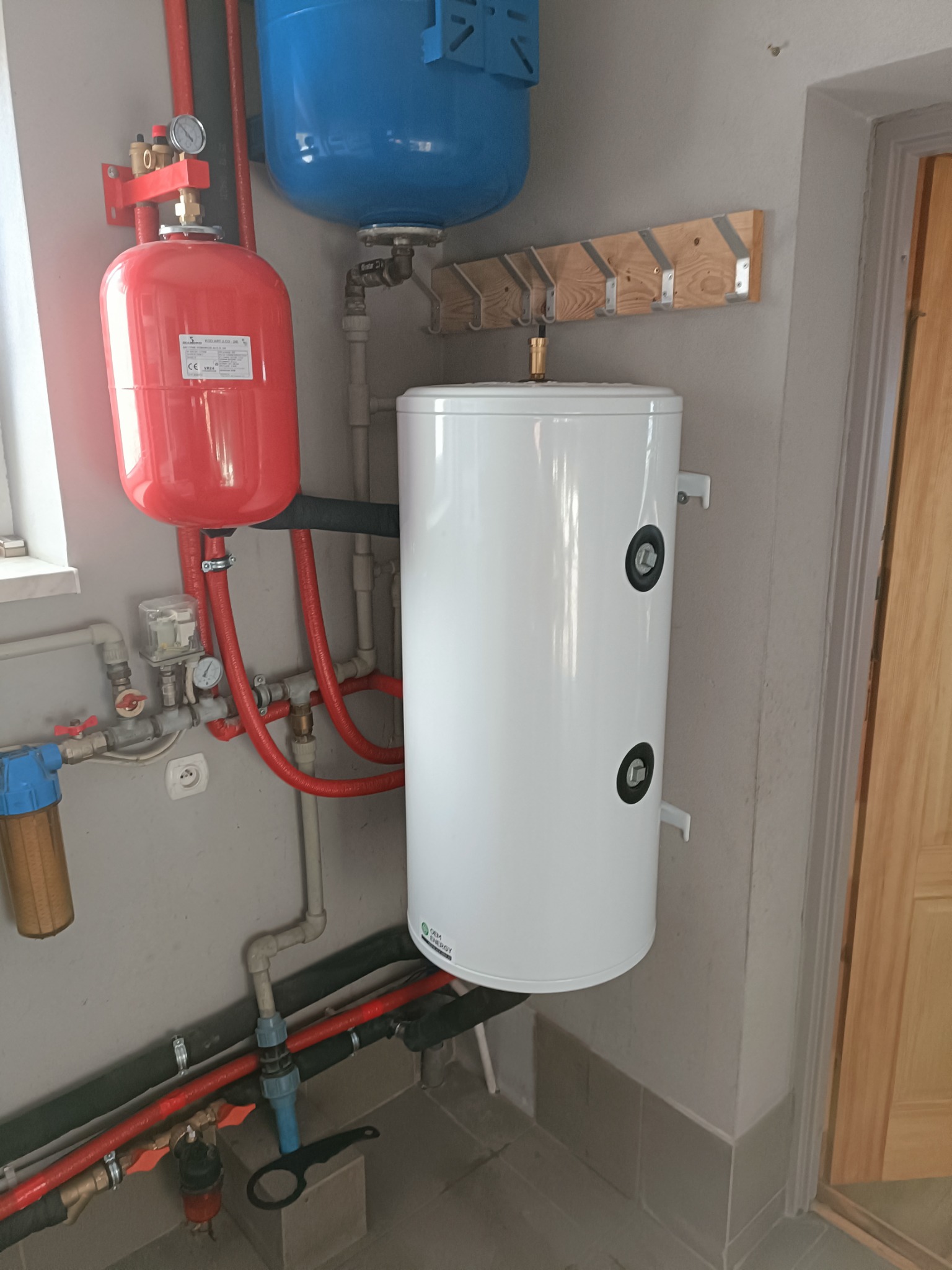 Biały, wiszący zasobnik ciepłej wody użytkowej z podłączonymi rurami miedzianymi i zaworami, zamontowany na szarej ścianie w kotłowni, obok czerwonego naczynia przeponowego i niebieskiego zbiornika...
