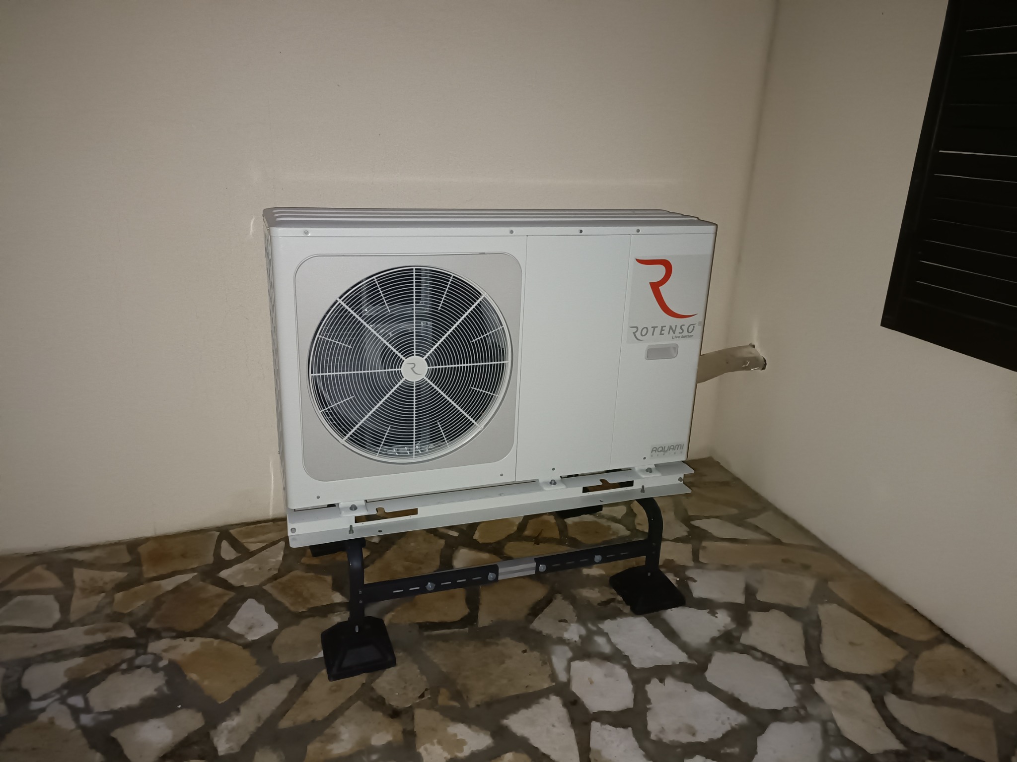 Biała pompa ciepła Rotenso Aquami, zamontowana na czarnym stelażu, na tle beżowej ściany i kamiennej podłogi w Rzeszowie.