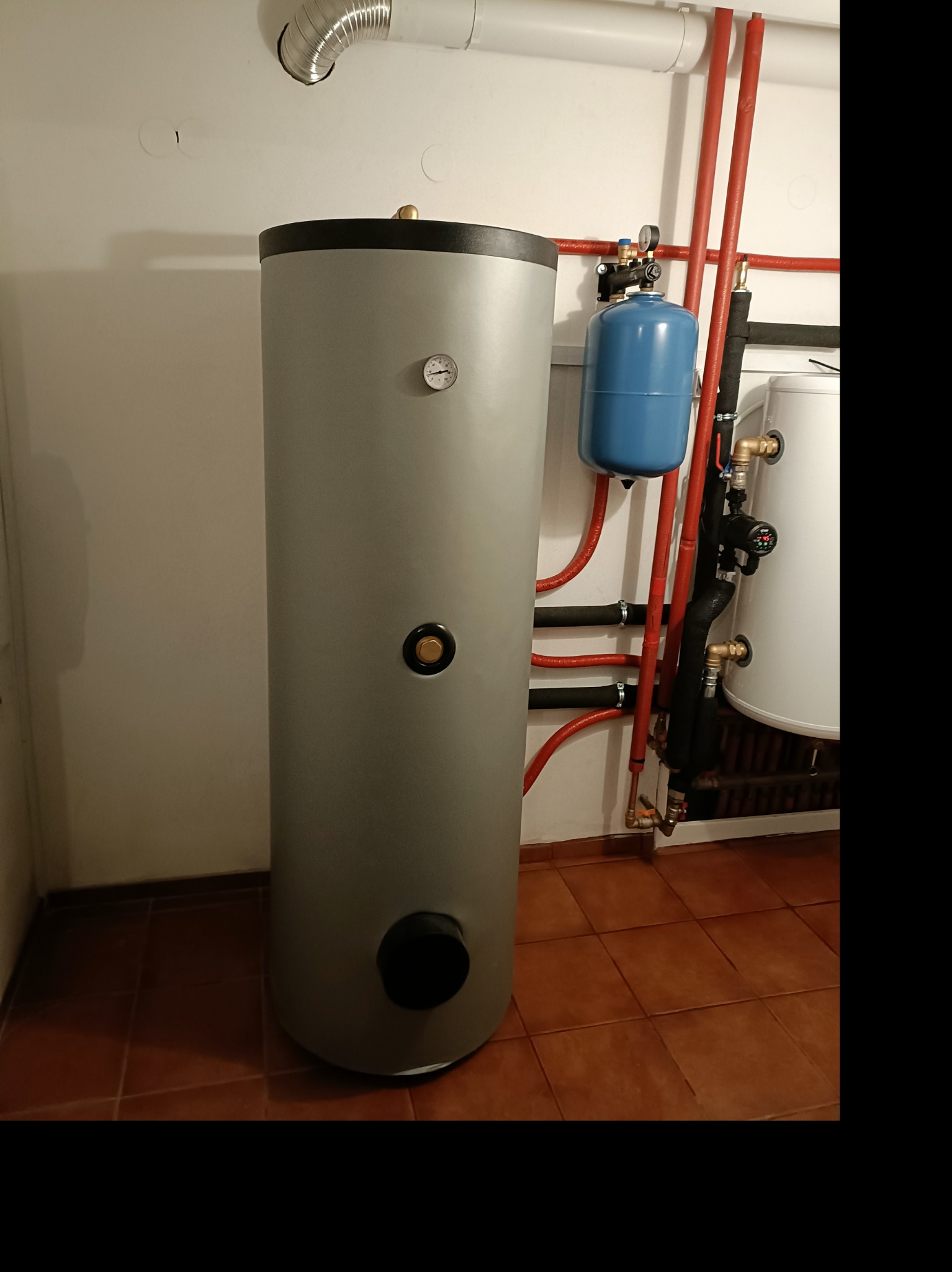 Szary, pionowy zbiornik na wodę z termometrem i czarnym otworem w dolnej części, obok niebieski zbiornik wyrównawczy i instalacja z czerwonych rur, na brązowej podłodze z płytek.