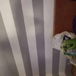 Ściana pomalowana w szerokie, pionowe pasy na szaro i biało, widoczne worki z farbą i narzędzia malarskie, sugerujące świeżo zakończone prace.